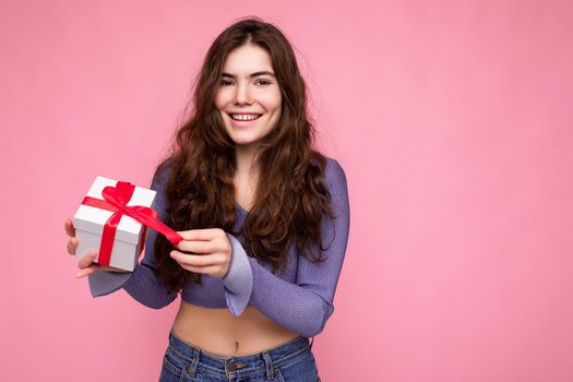 Shot of charming positive smiling brunette curly young woman isolated over pink background wall wearing purple blouse holding white gift box with red ribbon and unboxing present looking at camera.