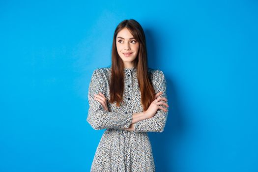 Smiling woman in romantic dress cross arms on chest and looking left at logo, standing against blue background.