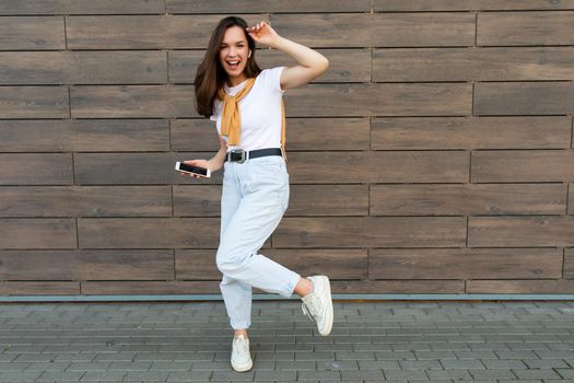 Full length body size photo of pretty brunet young happy woman wearing casual clothes and listening to music via wireless earphones standing in the street holding and using mobile phone looking at camera.Copy space
