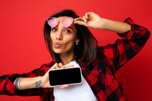 Photo of beautiful smiling young woman good looking wearing casual stylish red shirt white t-shirt and red sunglasses standing isolated on red background with copy space holding smartphone showing phone in hand with empty screen display for mockup looking at camera and giving kiss.