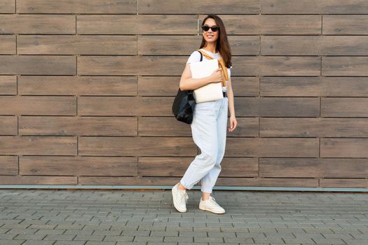 Full length photo shot of young charming beautifyl brunet woman going in the street near brown wall looking to side in white t-shirt and light blue jeans white sneakers with black female bag holding computer laptop and listening to music with wireless headphones.