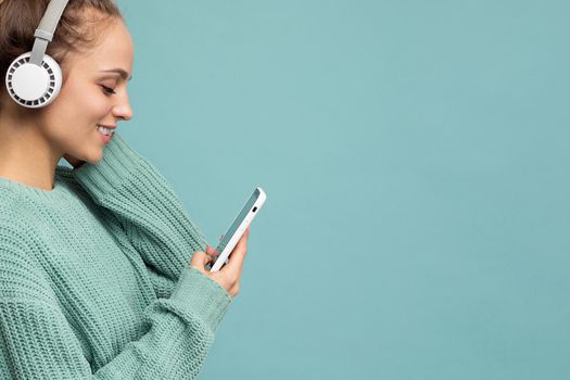 Closeup photo of beautiful joyful smiling young woman wearing stylish casual clothes isolated over background wall holding and using mobile phone wearing white bluetooth headphones listening to music and having fun looking to the side. copy space