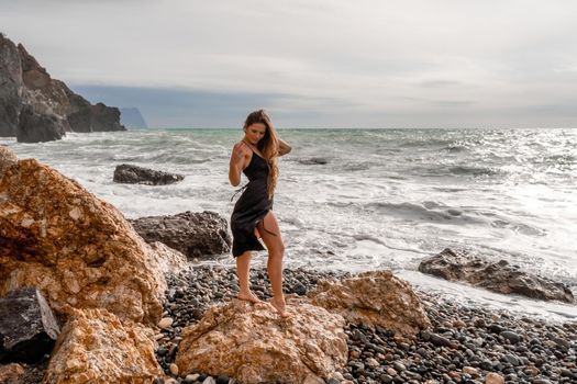 A beautiful girl in a black dress stands on a rock, big waves with white foam. A cloudy stormy day at sea, with clouds and big waves hitting the rocks