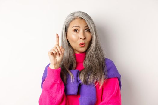 Close up of elderly asian woman in stylish hipster clothes, raising finger and looking up, having an idea, standing against white background.