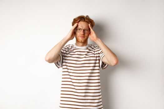 Image of young man with red hair and glasses touching head, frowning from painful migraine, having headache, standing over white background.