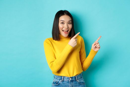 Shopping concept. Beautiful chinese girl in yellow sweater pointing fingers at upper right corner logo banner, smiling amused, standing over blue background.