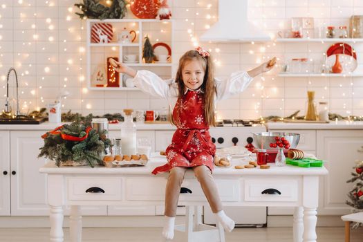 A happy little girl in the Christmas kitchen is sitting on the table with cookies in her hands.