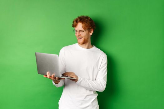 Smiling young man with red hair, wearing glasses, working on laptop and smiling, standing over green background.