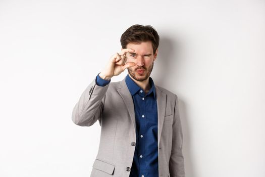 Businessman trying to see something tiny, showing small thing size with fingers, standing in suit on white background.