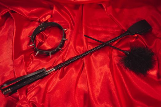 Bondage, kinky adult sex games, kink and BDSM lifestyle concept with a whip, feather stick, collar on red silk with copy space