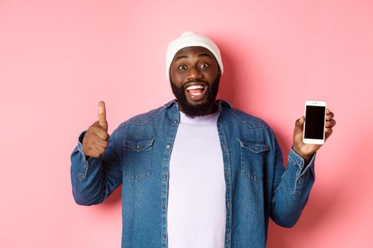 Online shopping and technology concept. Happy african-american hipster man showing thumbs-up and mobile screen, praising app, standing over pink background.