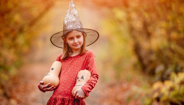 Little girl in witch hat holding decorated pumpkins for halloween on colorful autumn nature background