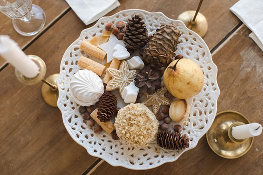Close up view of white patterned plate with pine cones and candies on wooden table decorated with candles and wine glass