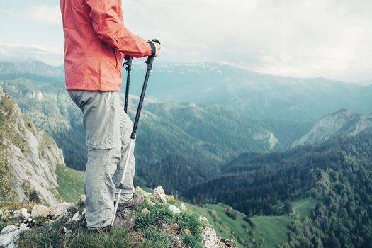 Unrecognizable female explorer with trekking poles standing on peak of mountain in summer.