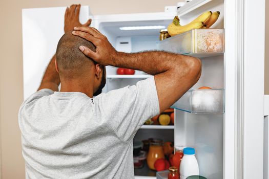 African american man standing in front of open fridge and choosing food, back view