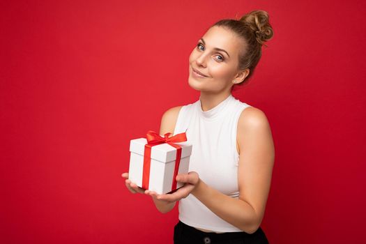Pretty happy young blonde female person isolated over red background wall wearing white top holding gift box and looking at camera. Empty space
