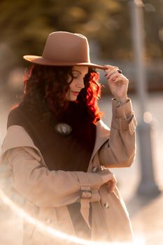 Outdoor fashion portrait of young elegant fashionable brunette woman, model in stylish hat, choker and light raincoat posing at sunset in European city