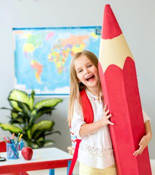 Little smiling blond girl with red bag holding huge red decorative pencil on her shoulder in the school classroom