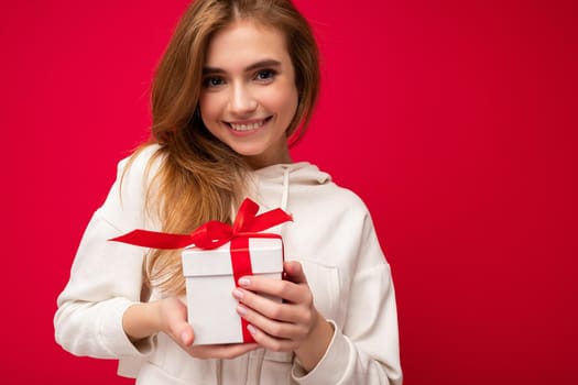 Photo shot of attractive happy smiling cute blonde young female person isolated over red background wall wearing white hoodie holding white gift box with red ribbon and looking at camera. Free space