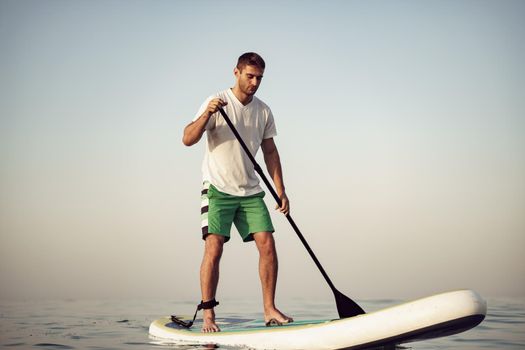 Young man in t-shirt and shorts floating on SUP board at sea