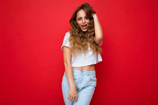 Young beautiful blond woman wearing white t-shirt looking at camera. Positive female shows facial sincere emotions. Funny model isolated on red wall with empty space.