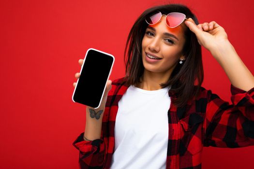 Photo of beautiful smiling young woman good looking wearing casual stylish red shirt white t-shirt and red sunglasses standing isolated on red background with copy space holding smartphone showing phone in hand with empty screen display for mockup looking at camera.