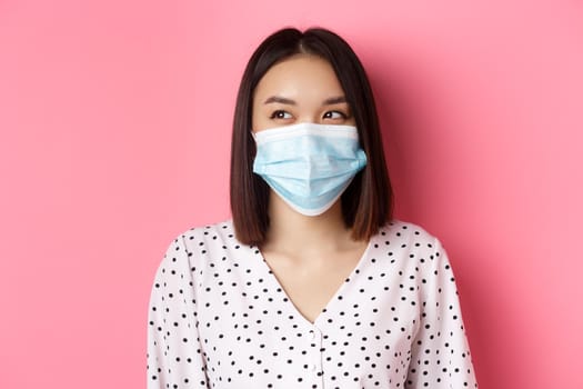 Covid-19, pandemic and lifestyle concept. Beautiful asian female model in medical mask laughing, smiling and looking left at copy space, standing over pink background.