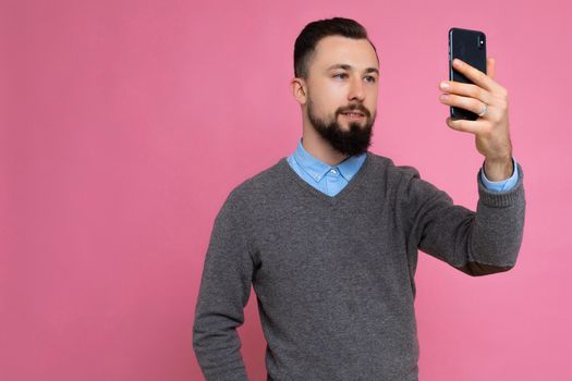 Handsome young man wearing casual stylish clothes standing isolated over background wall holding smartphone taking selfie photo looking at mobile phone screen display