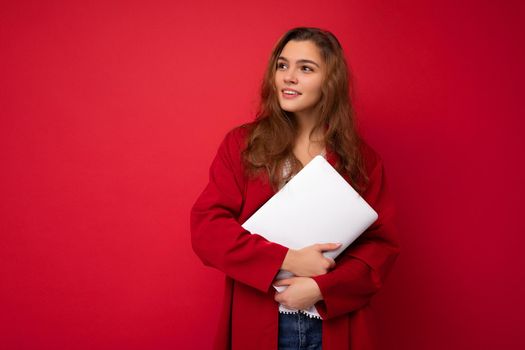 Charming mysterious young curly lady holding netbook wearing red cardigan and white blouse looking to the side isolated over red background. Copy space