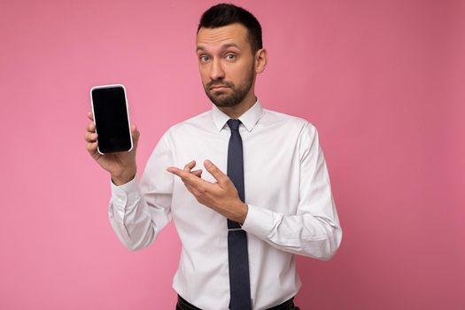 Self-confident handsome good looking man wearing casual white shirt and tie isolated on pink background with empty space holding in hand and showing mobile phone with empty screen for mockup looking at camera.