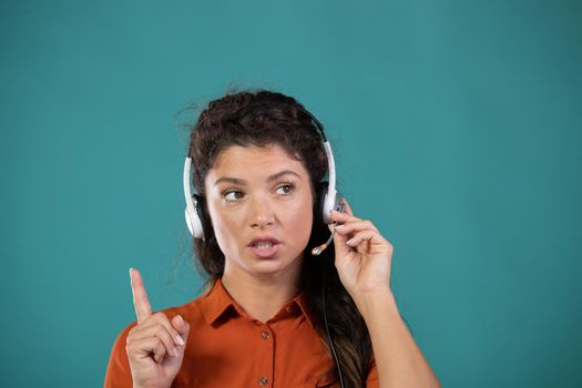 Pretty young woman operator with headphones holding forefinger up, talking and solving problem with customer, on blue background in studio