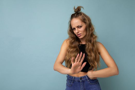 Young sad upset beautiful winsome blonde curly woman with sincere emotions wearing stylish black top isolated over blue background with copy space and showing no gesture.