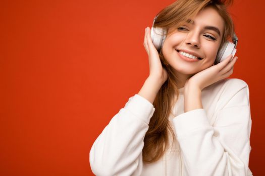Photo of beautiful sexy happy smiling young blonde woman wearing white hoodie isolated on colourful background wearing white wireless bluetooth headphones listening to cool music and enjoying looking to the side.