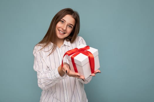 Beautiful happy young dark blonde woman isolated over colourful background wall wearing stylish casual clothes holding gift box and looking at camera. Copy space, mockup
