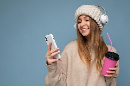 Attractive happy smiling young blonde woman wearing beige sweater and beige hat white headphones isolated over blue background holding in hand and using mobile phone reading news drinking beverage and listening to music looking at gadjet display.