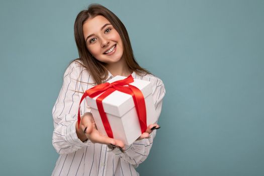 Shot of attractive positive smiling young dark blonde woman isolated over colourful background wall wearing everyday trendy outfit holding gift box and looking at camera. Empty space