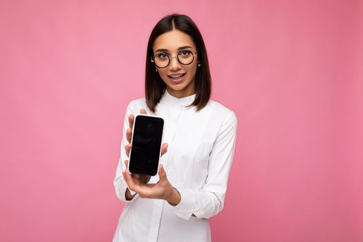 charming smiling young brunette woman wearing white blouse and optical glasses standing isolated over pink background showing mobile phone with empty screen for mockup looking at camera.