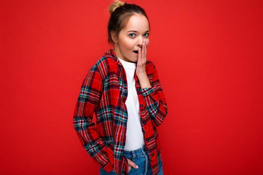 Attractive amazed young blonde woman standing isolated over red wall wearing casual red shirt and white t-shirt looking at camera. Copy space