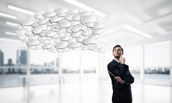 Portrait of young thoughtful man holding paintbrush in his hand and looking away while standing with chatting cloud inside modern office view on background.