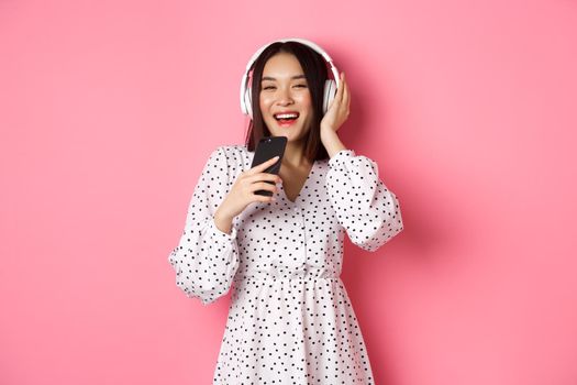 Beautiful smiling asian woman singing song in smartphone microphone, playing karaoke app and using headphones, standing over pink background.