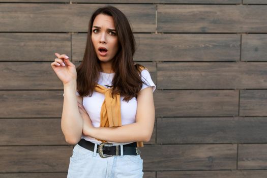 Portrait of sad upset sorrowful dissatisfied and asking young brunet woman wearing casual white t-shirt with yellow sweater poising near brown wall in the street.
