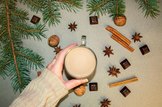 Hot Christmas Drink Cocoa Coffee or Chocolate with Milk in a Small Transparent Cup. Cup in Hand. Fir Tree Branch, Nuts, Cinnamon Sticks Star Anise on a Grey Background. Top View Winter time. New Year