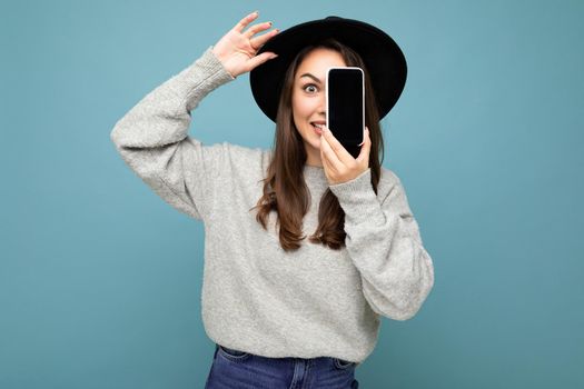 Pretty young smiling woman wearing black hat and grey sweater holding phone looking at camera isolated on background. cutout