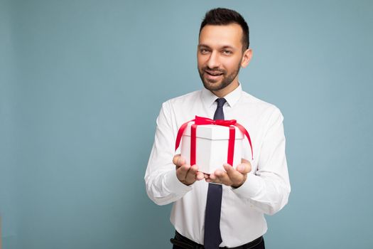 Handsome positive brunette young unshaven businessman male peson with beard isolated over blue background wall wearing white shirt and tie holding white gift box with red ribbon and looking at camera. Free space