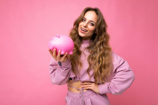 Portrait of happy positive smiling young beautiful attractive woman with wavy long blonde hair with sincere emotions wearing stylish pink hoodie isolated over pink background with copy space and holding pink piggy box. Money box concept.