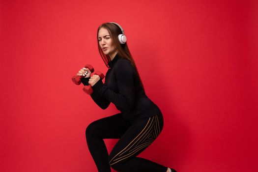 Beautiful happy smiling young brunet woman wearing black sport clothes isolated on red background wall doing fitness using dumbbells wearing white bluetooth headsets listening to music looking at camera.