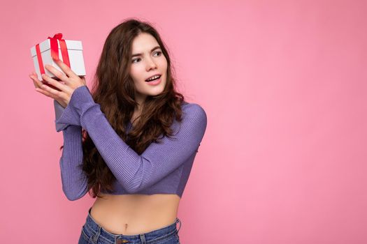 Photo shot of beautiful happy brunette curly young woman isolated over pink background wall wearing purple blouse holding white gift box with red ribbon and looking to the side. Copy space