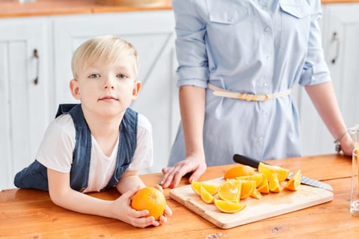 Healthy food, fresh fruit, juicy oranges. Mother and son are smiling while having a breakfast in kitchen. Bright morning in the kitchen