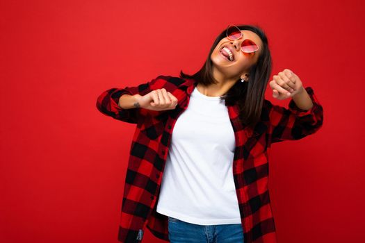 Portrait of young happy positive beautiful brunette woman with sincere emotions wearing white t-shirt, stylish red check shirt and red sunglasses isolated on red background with copy space and having fun.