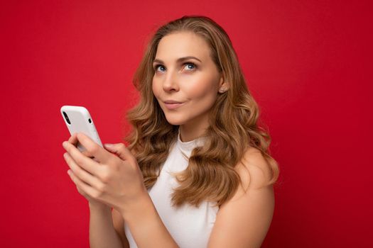 Closeup of thoughtful happy pretty young blonde woman wearing white t-shirt isolated over red background using smartphone and chatting via mobile phone looking to the side and dreaming.